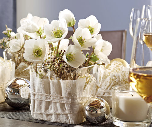Christrose als Schnittblume in einem mit Wachs gefertigten Serviettengefäß festlich mit Christbaumkugeln und Kerzen auf dem Tisch dekoriert
